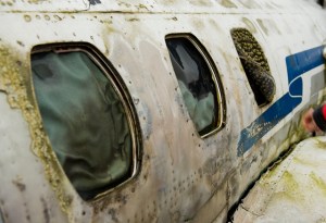 Burnt aircraft fuselage showing the rounded window frames, distorted plastic inner window (first two) and the charcoaled outer window pane (3rd window) (Photo R De Crespigny)